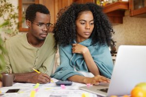 Young African-American family doing paperwork together, sitting at kitchen table with piles of papers, looking at laptop screen with serious and concentrated expressions, paying utility bills online