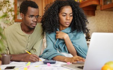 Young African-American family doing paperwork together, sitting at kitchen table with piles of papers, looking at laptop screen with serious and concentrated expressions, paying utility bills online