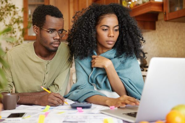 Young African-American family doing paperwork together, sitting at kitchen table with piles of papers, looking at laptop screen with serious and concentrated expressions, paying utility bills online